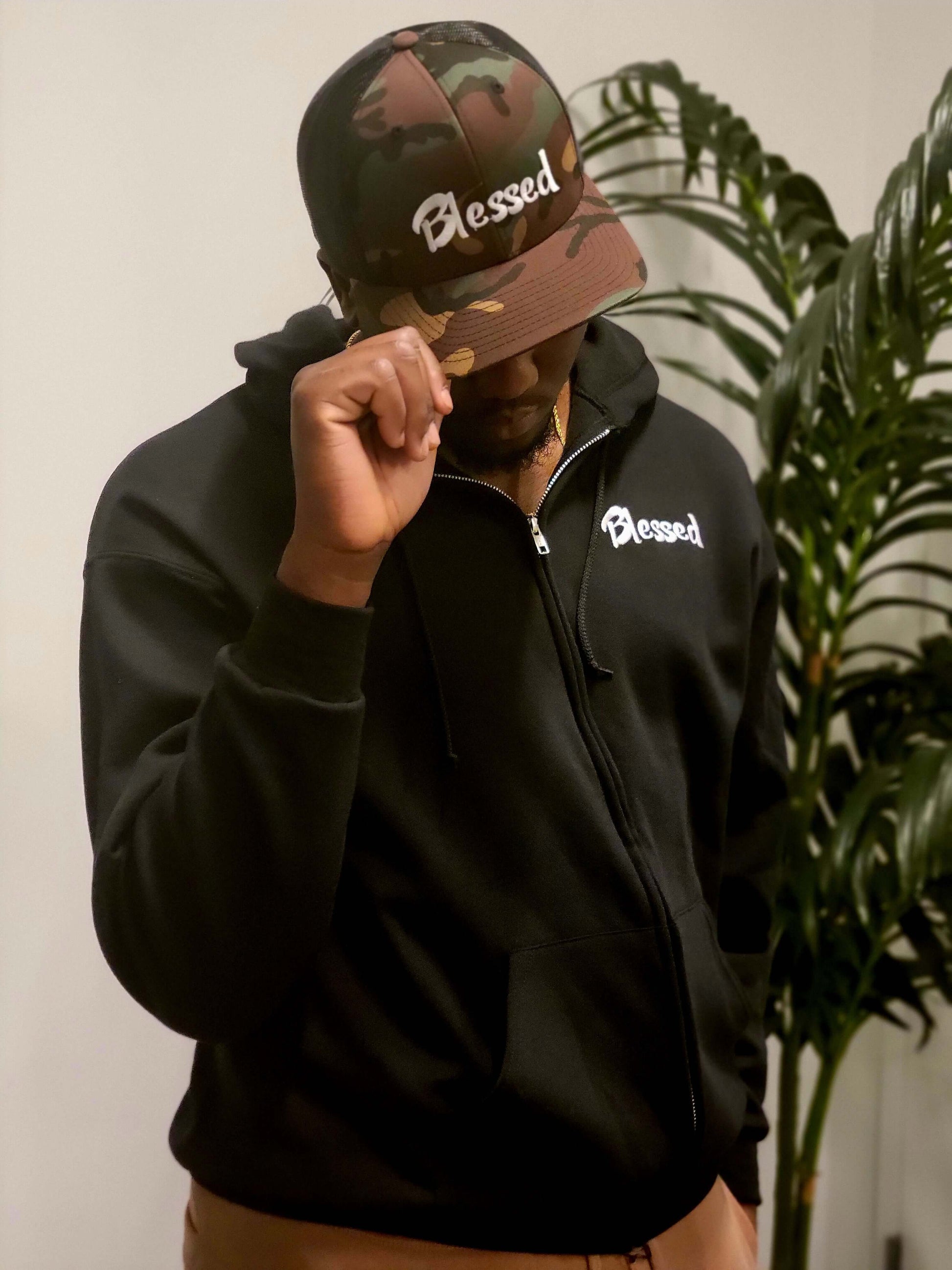 A man wearing a full-zip hooded sweatshirt made with a 50/50 cotton-polyester with embroidered "Blessed" text in white font with a camouflage cap written "blessed" in white font.