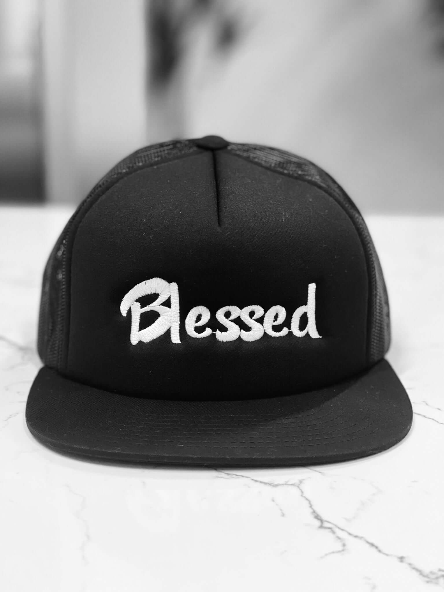 Black trucker hat made of 64/35 polyester/cotton with the word "Blessed" embroidered in white.