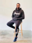 A man wearing a Black Unisex Hoodie and black jogger pants with the word "blessed" written in white font.